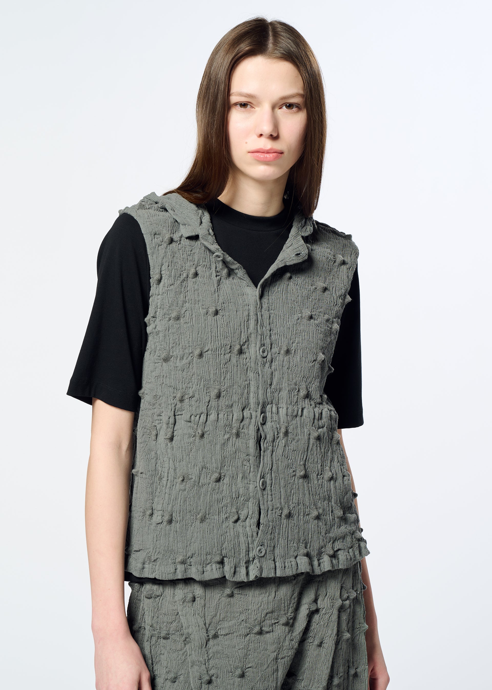 Zoomed view A person with straight brown hair wears the HaaT KYO CHIJIMI SQUARE EMB vest—a textured gray sleeveless piece featuring subtle embroidered square patterns—over a black shirt. They stand against a plain light background, looking at the camera with a neutral expression.