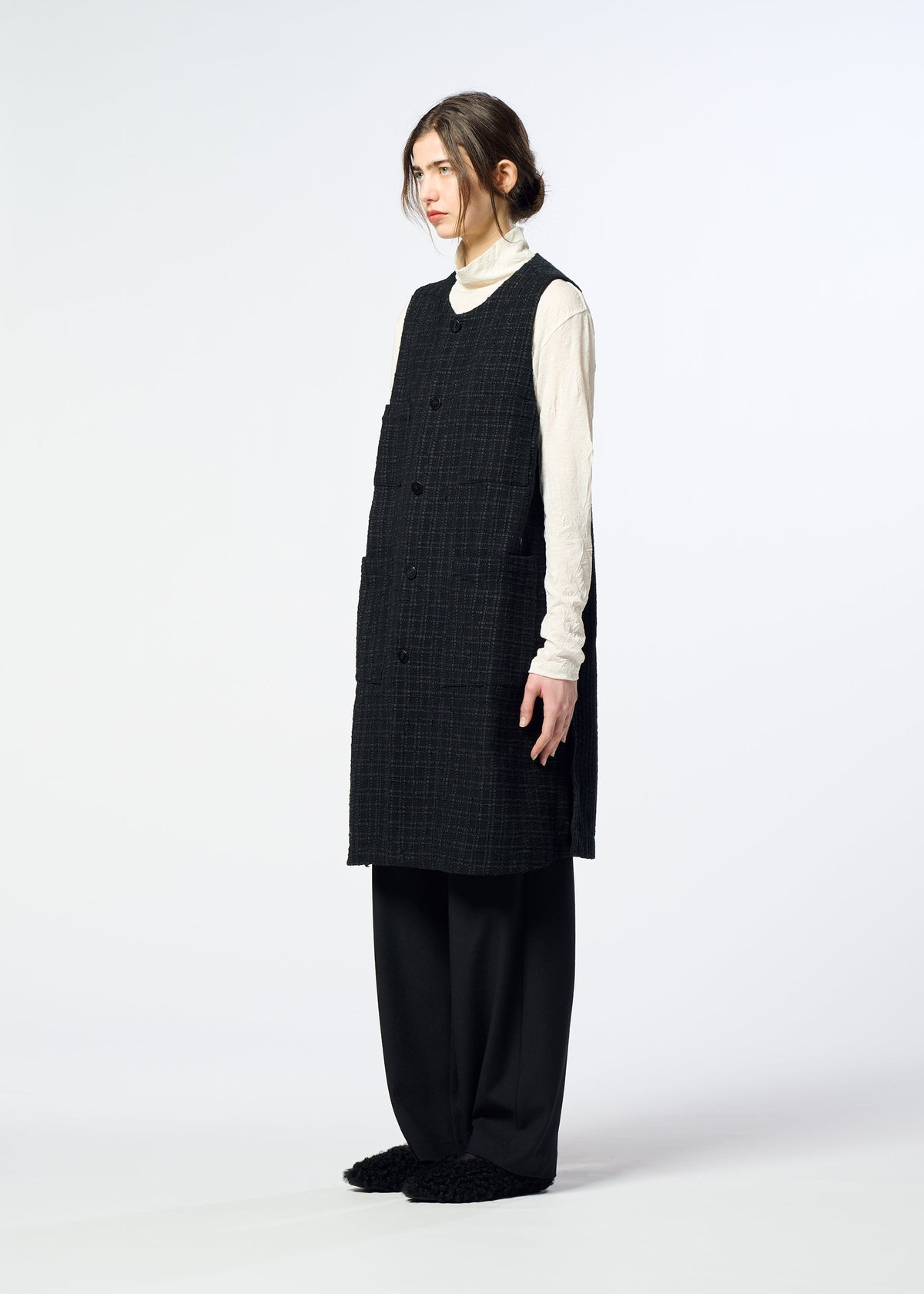 Zoomed view Person standing sideways in the SAKIORI long, sleeveless black woven vest by HaaT, layered over a cream turtleneck with loose black pants and dark slippers; hair tied back in a low bun against a plain background.