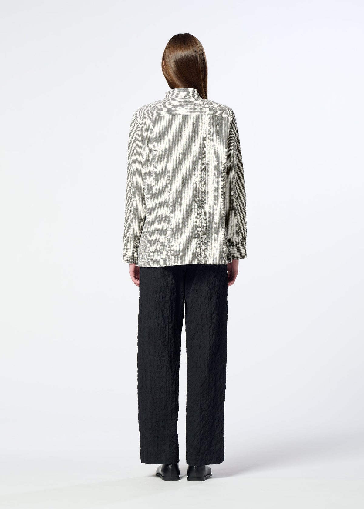 A person stands with their back to the camera, wearing HaaT’s RIPPLE STRIPE black seersucker pants featuring a subtle striped pattern, paired with a light textured jacket and black shoes, set against a plain white background. view 8 of 8