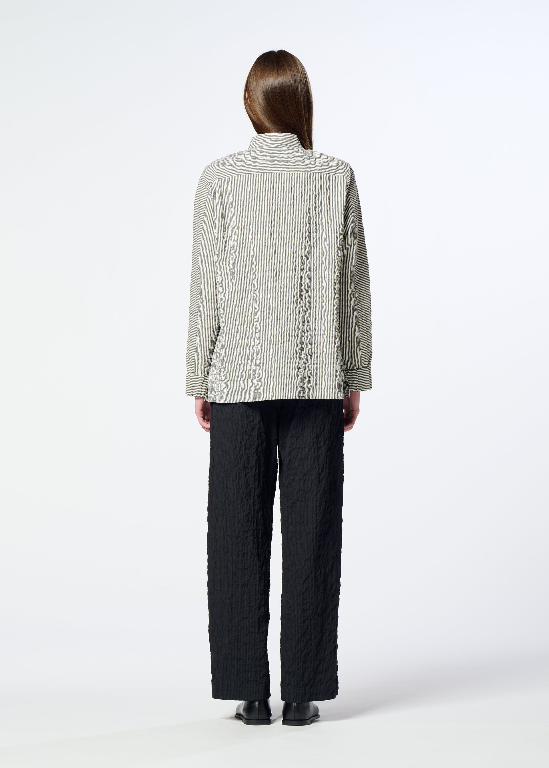 Zoomed view A person stands with their back to the camera, wearing HaaT’s RIPPLE STRIPE black seersucker pants featuring a subtle striped pattern, paired with a light textured jacket and black shoes, set against a plain white background.