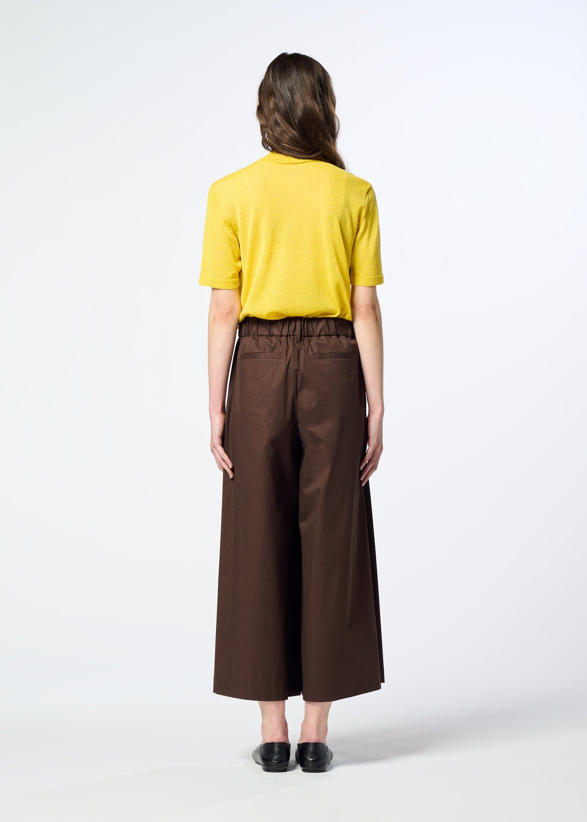A person stands facing away, wearing a yellow short-sleeve shirt crafted from organic cotton, HaaT’s COTTON STRETCH BOTTOM in brown, and black shoes, against a plain light background. view 8 of 8