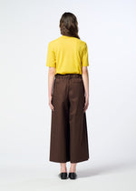 A person stands facing away, wearing a yellow short-sleeve shirt crafted from organic cotton, HaaT’s COTTON STRETCH BOTTOM in brown, and black shoes, against a plain light background.