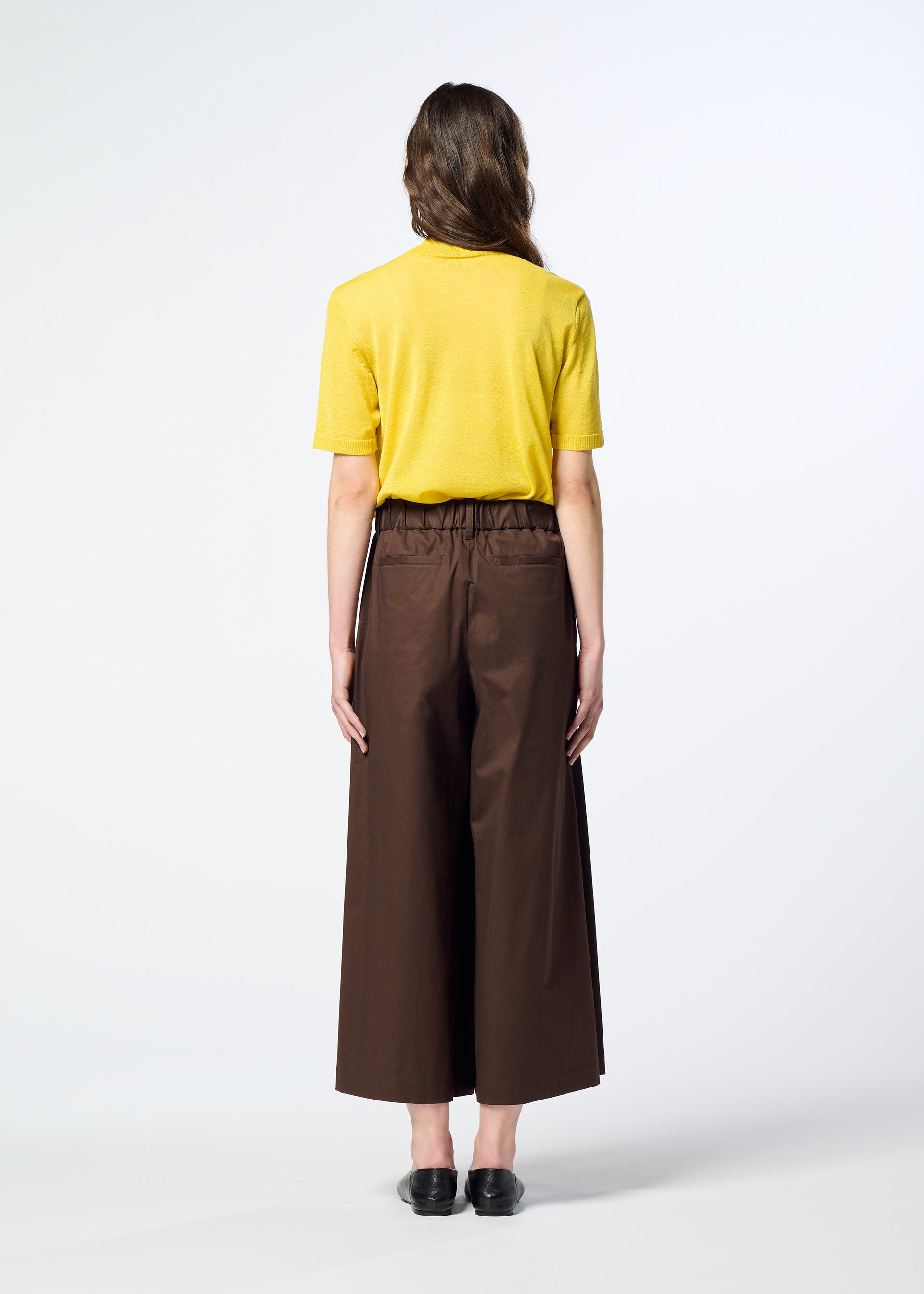 Zoomed view A person stands facing away, wearing a yellow short-sleeve shirt crafted from organic cotton, HaaT’s COTTON STRETCH BOTTOM in brown, and black shoes, against a plain light background.