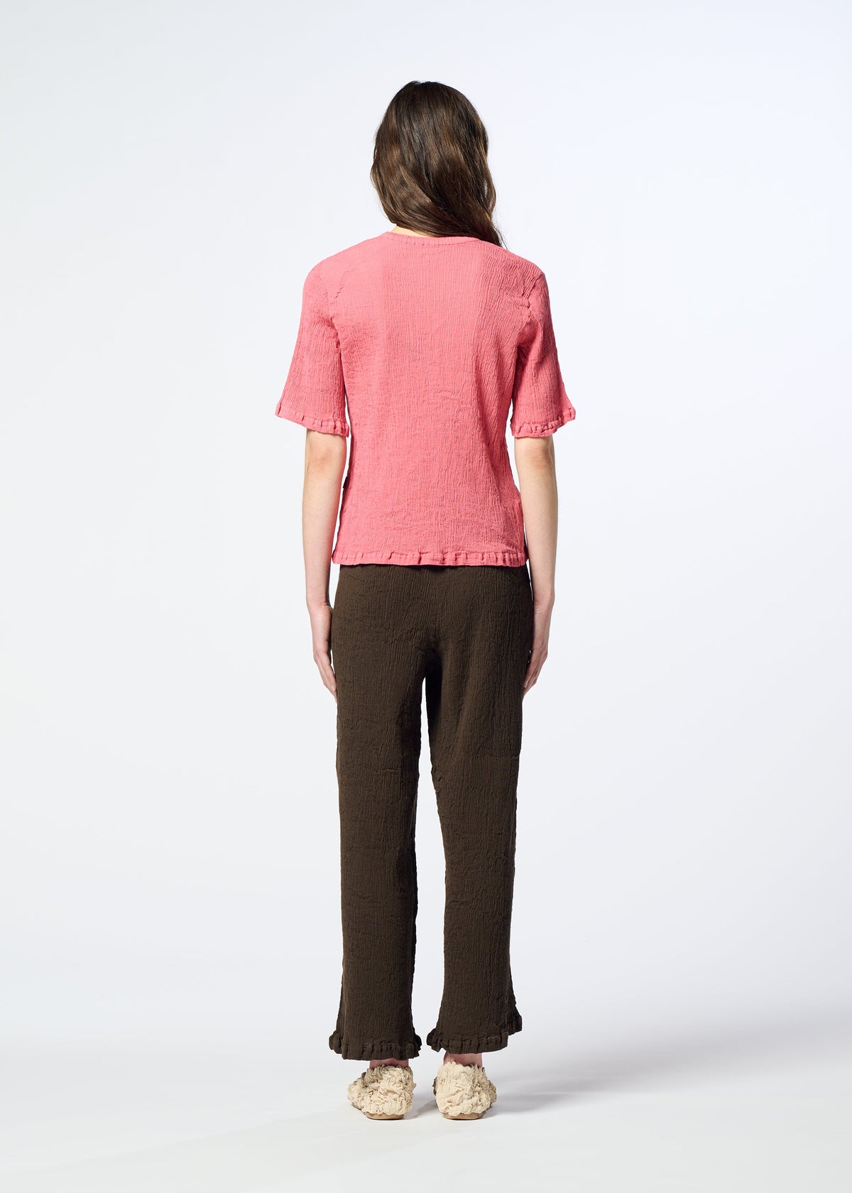 A person stands facing away, wearing a pink short-sleeve textured top, HaaT’s KYO CHIJIMI AUGUST pants in dark brown featuring a distinctive crinkled texture and ruffled hems, paired with beige shoes. The background is plain and well-lit. view 5 of 5