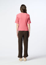 A person stands facing away, wearing a pink short-sleeve textured top, HaaT’s KYO CHIJIMI AUGUST pants in dark brown featuring a distinctive crinkled texture and ruffled hems, paired with beige shoes. The background is plain and well-lit.