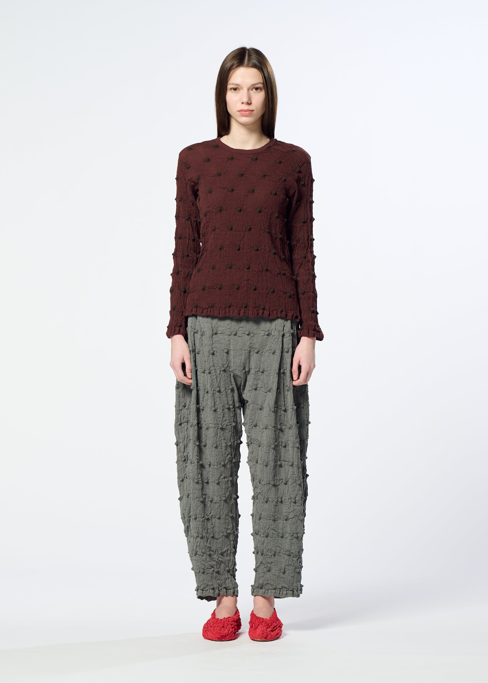 Zoomed view A woman stands facing forward against a plain white background, wearing a textured maroon long-sleeve top, gray KYO CHIJIMI SQUARE EMB pants by HaaT featuring a distinctive crinkled texture, and red textured shoes.