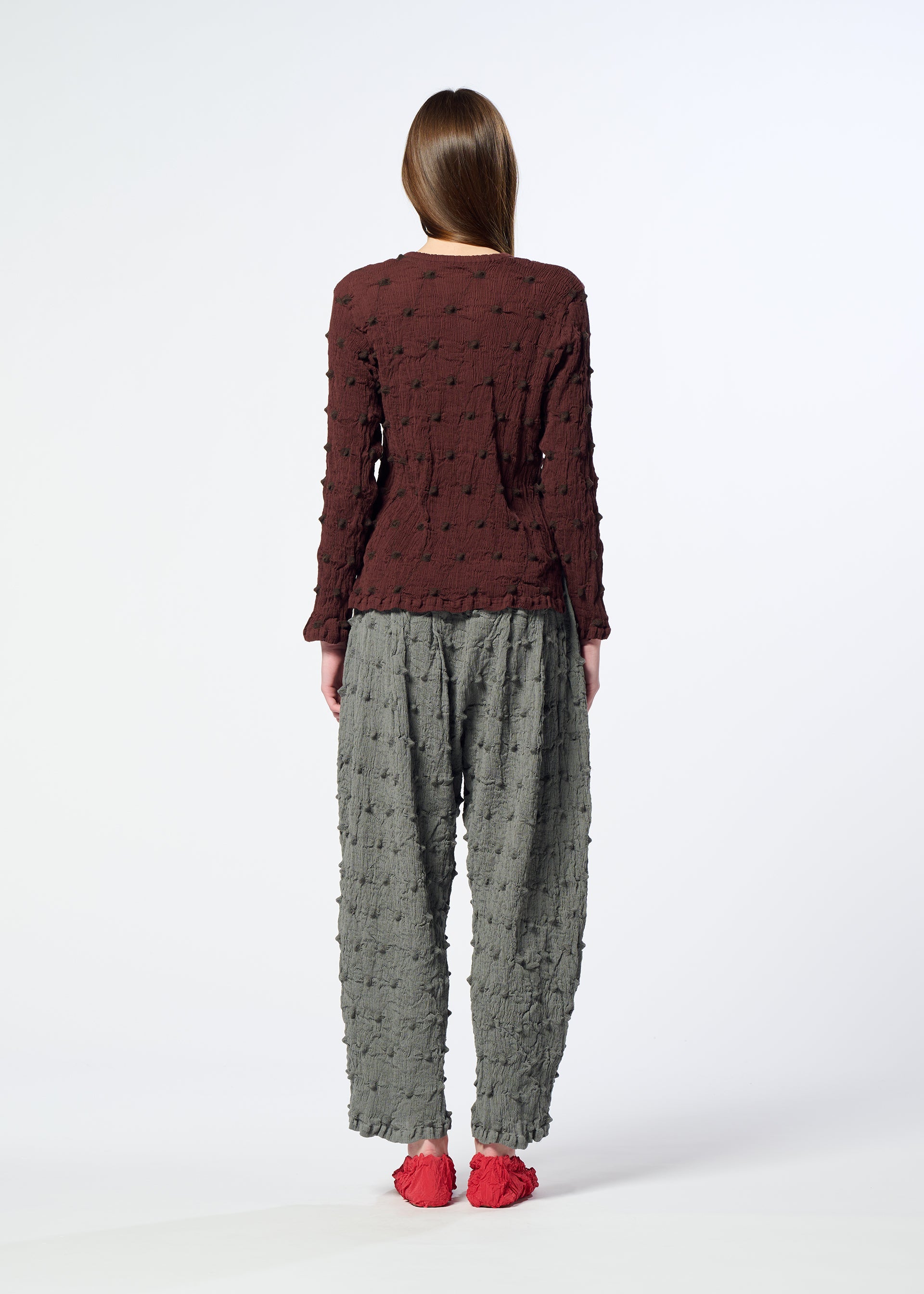 Zoomed view A person with long brown hair stands facing away against a plain white background, wearing HaaT’s KYO CHIJIMI SQUARE EMB—a textured maroon top with subtle square-pattern embroidery—paired with loose crinkled gray pants and red shoes.