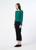 A person with straight brown hair faces left, wearing a textured green button-up top and HaaT's KYO CHIJIMI TSUCHIKABE pants—featuring a crepe-like texture and ruffled hems—with black shoes, set against a plain light background.