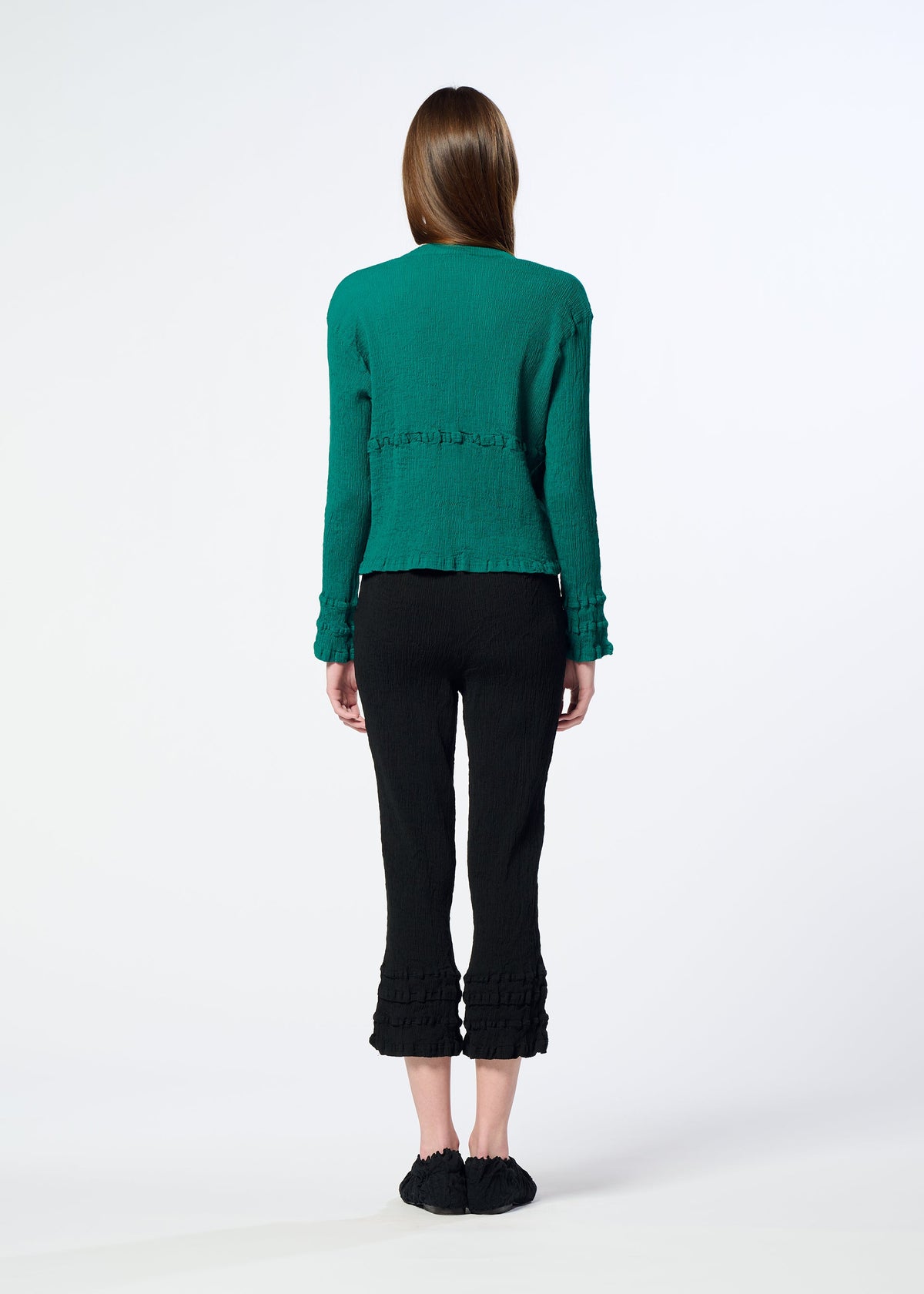 A woman faces away, wearing a textured green long-sleeve top and black HaaT KYO CHIJIMI TSUCHIKABE pants with ruffled hems, crafted from ultra-fine cotton for a crepe-like feel. Black slip-on shoes complete the look against a plain white background. view 9 of 9