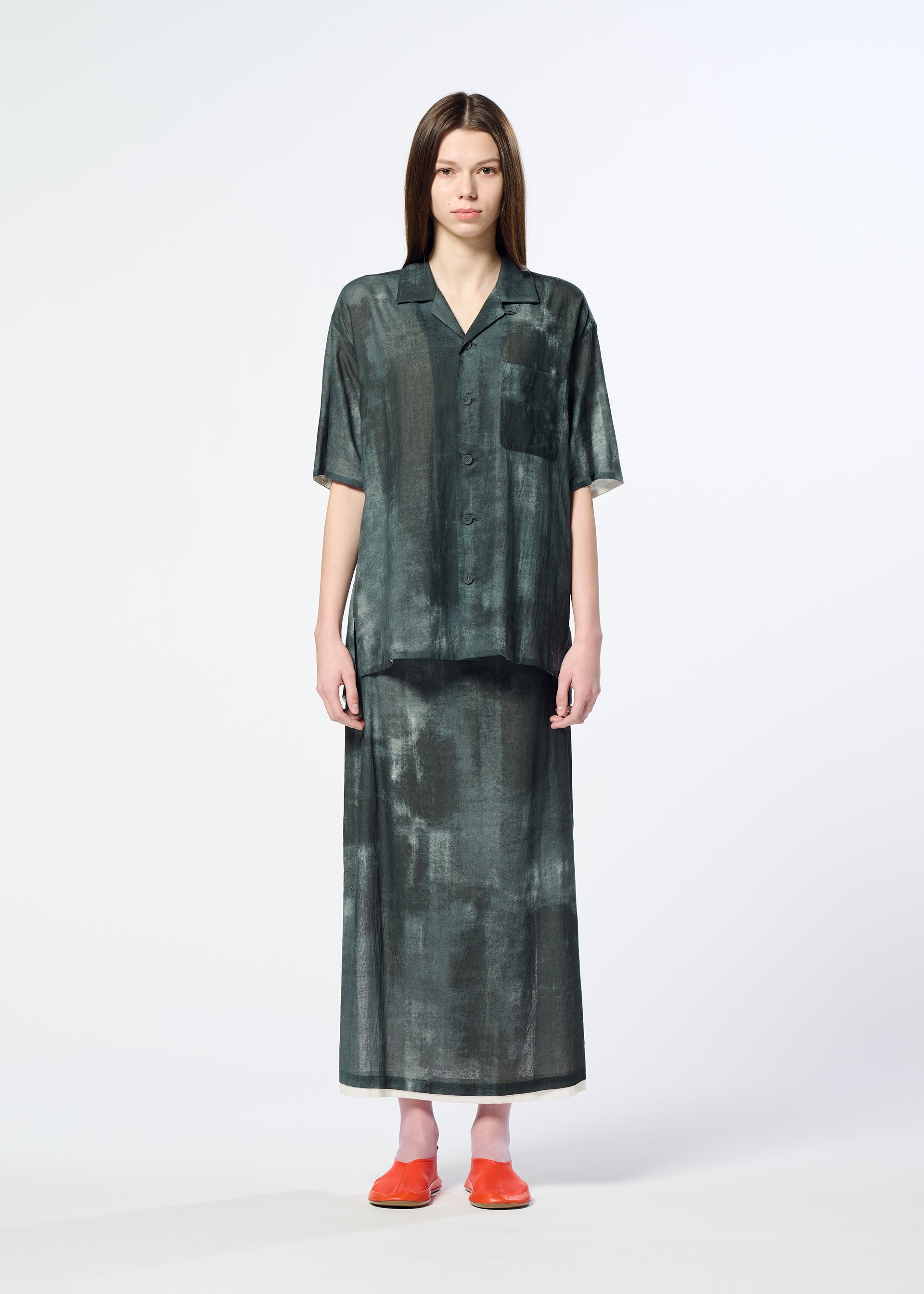 Zoomed view A woman stands against a plain background wearing HaaT's WALL WHITE HEM, a loose-fitting, dark green shirt paired with a matching Kyoto dyeing chiffon skirt and bright orange shoes.