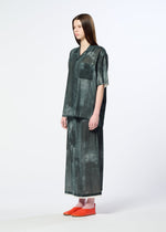 A woman with straight brown hair wears HaaT’s WALL WHITE HEM oversized dark green short-sleeve button-up shirt with chest pocket and a matching Nassen print long skirt, paired with orange slip-on shoes, against a plain background.