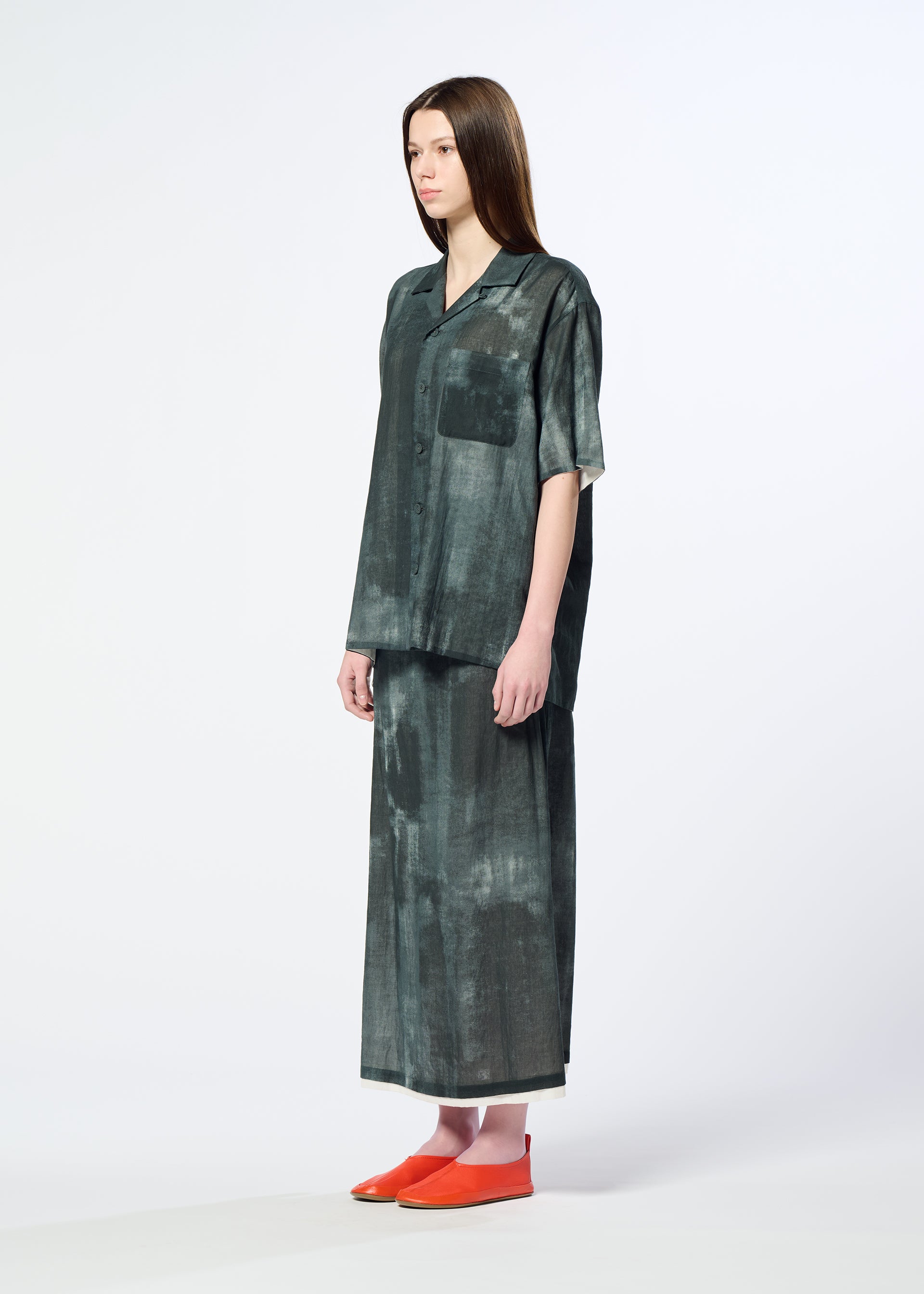 Zoomed view A woman with straight brown hair wears HaaT’s WALL WHITE HEM oversized dark green short-sleeve button-up shirt with chest pocket and a matching Nassen print long skirt, paired with orange slip-on shoes, against a plain background.