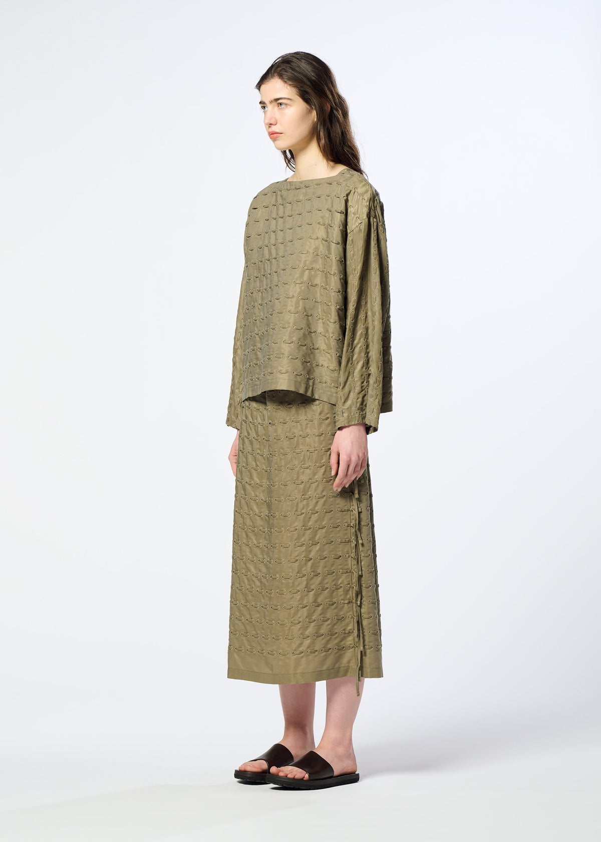 A woman stands in profile wearing HaaT’s HOLE & LINE set—an olive green, textured top paired with a matching long twill skirt featuring subtle eyelet embroidery—and black sandals, against a plain light background. view 7 of 8