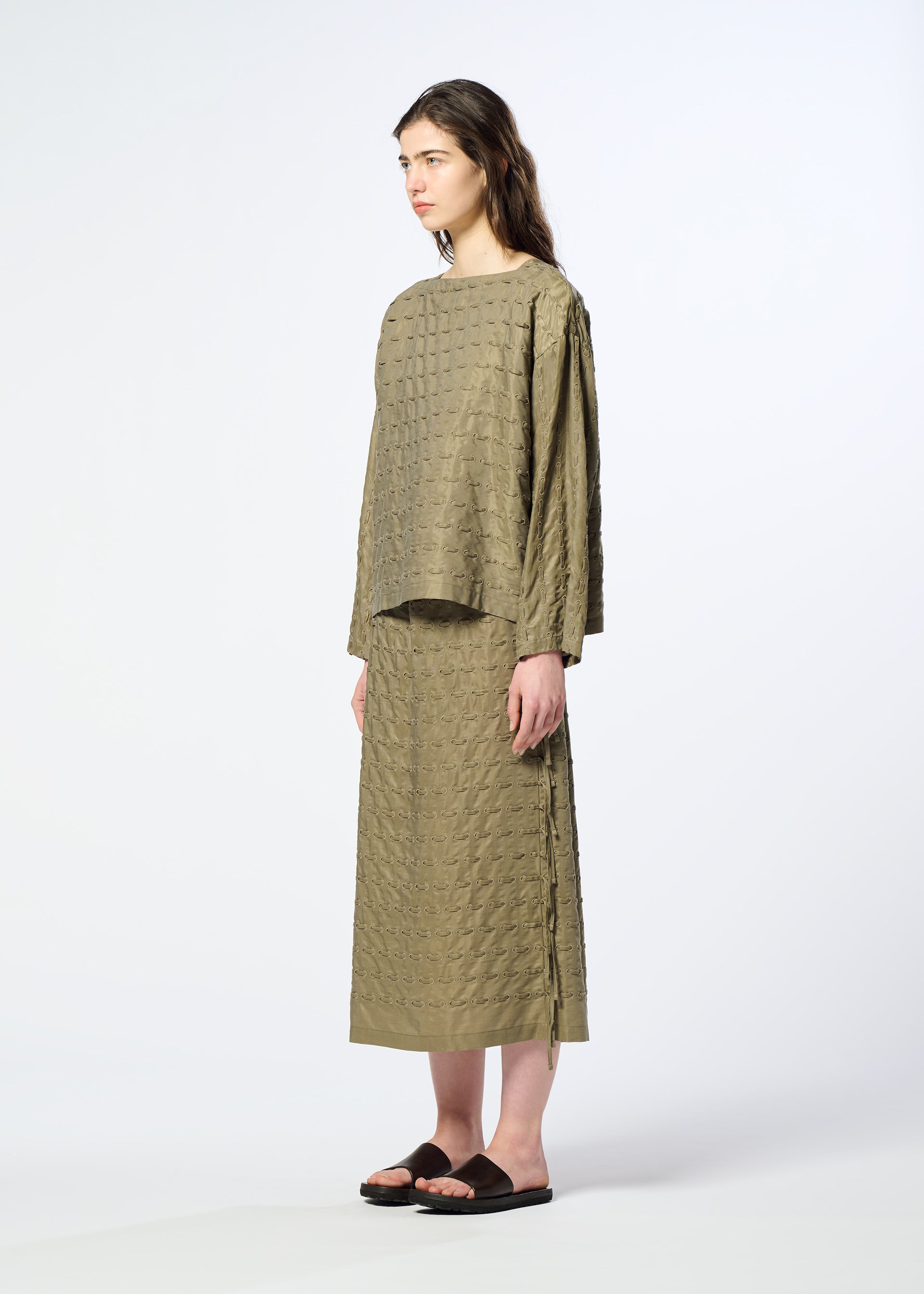 Zoomed view A woman stands in profile wearing HaaT’s HOLE & LINE set—an olive green, textured top paired with a matching long twill skirt featuring subtle eyelet embroidery—and black sandals, against a plain light background.
