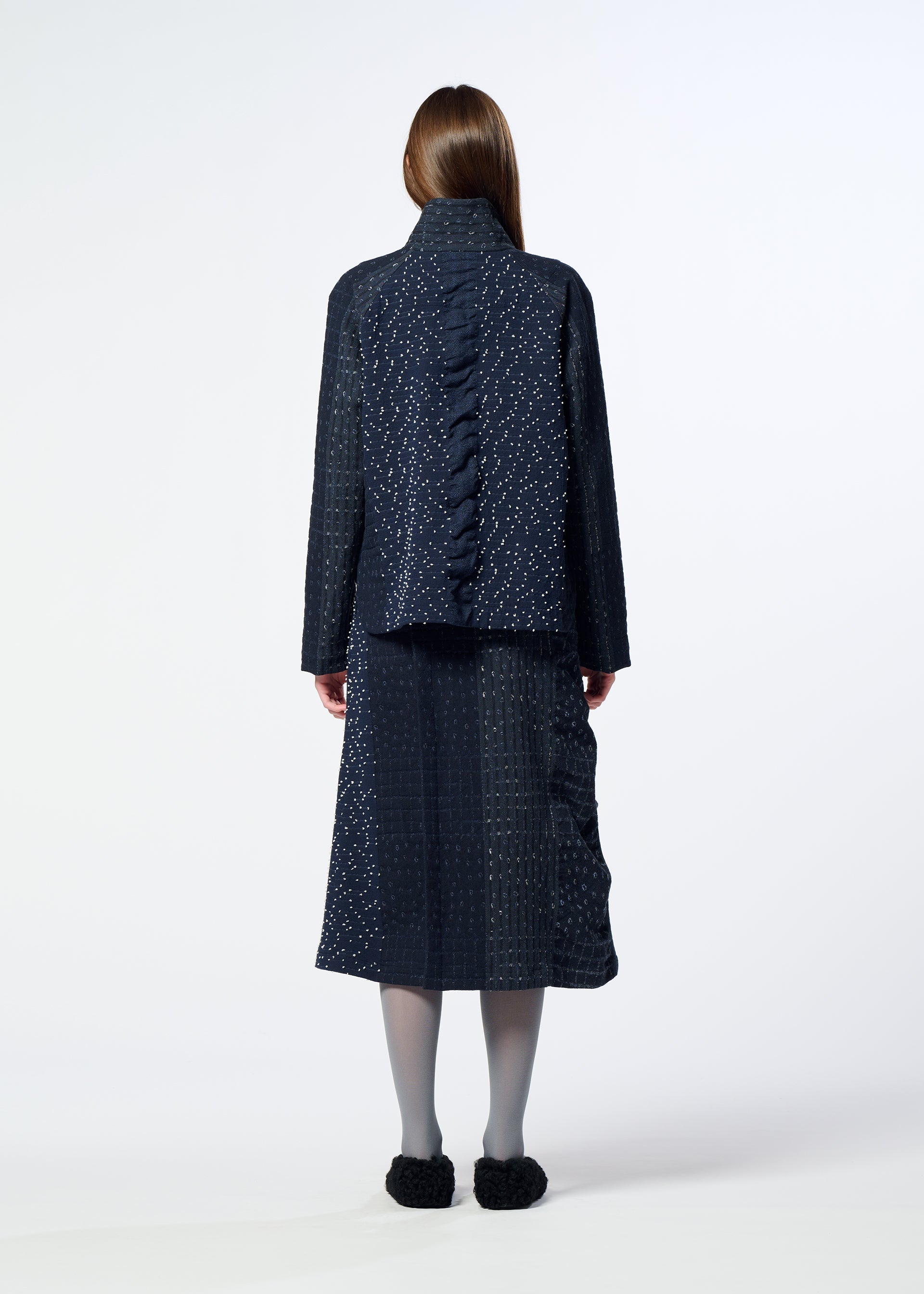 Zoomed view A person with long brown hair stands facing away, wearing HaaT’s COLLAGE STRETCH Ⅰ dark blue textured jacquard skirt and matching jacket with small white dots, paired with gray tights and black shoes against a plain white background.