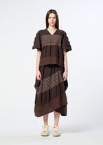 A woman stands facing forward against a plain white background, wearing HaaT’s COLLAGE STRETCH Ⅱ—a loose brown dress featuring a striped pattern and ruffled details that create a voluminous silhouette—paired with cream sneakers.