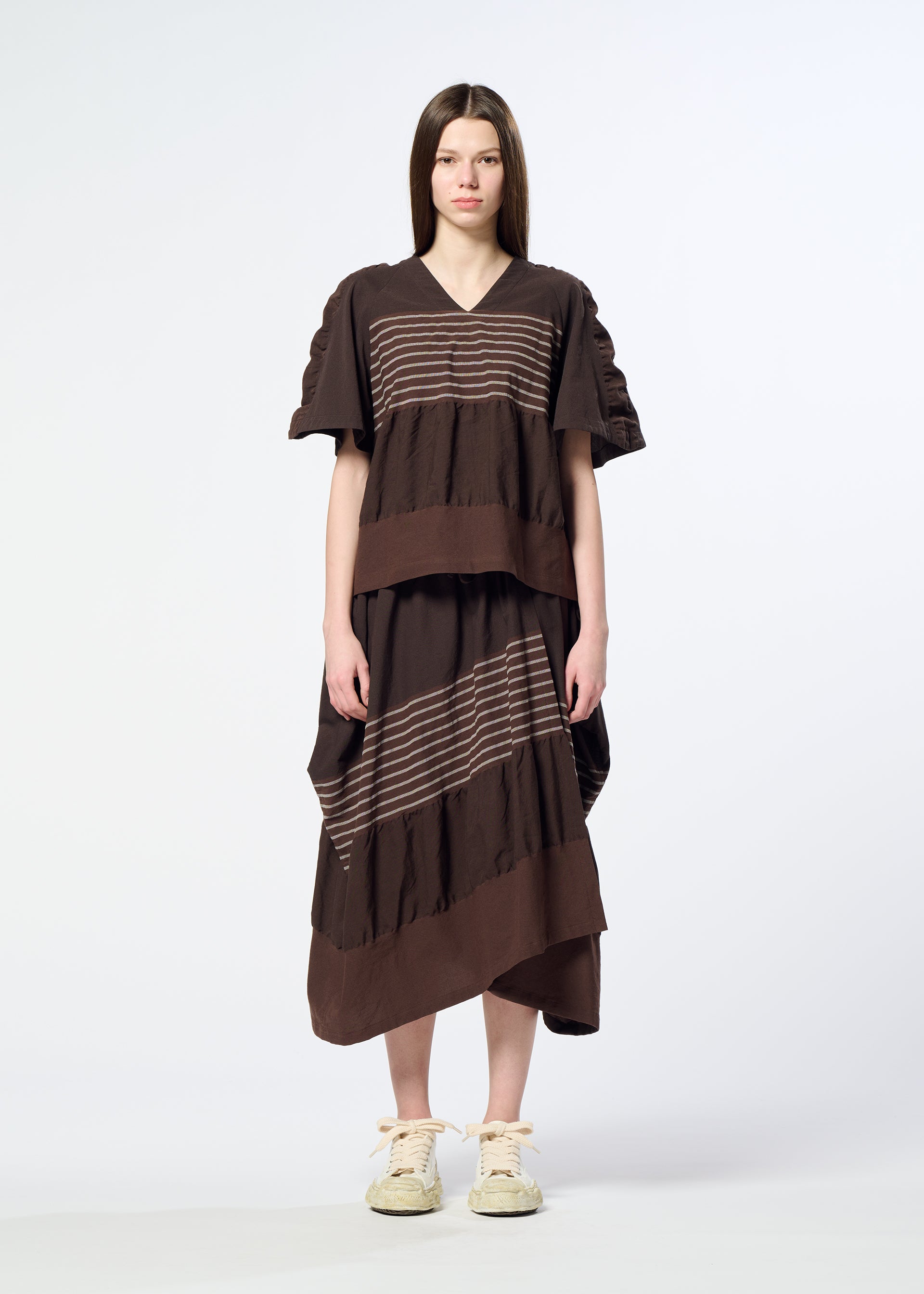 Zoomed view A woman stands facing forward against a plain white background, wearing HaaT’s COLLAGE STRETCH Ⅱ—a loose brown dress featuring a striped pattern and ruffled details that create a voluminous silhouette—paired with cream sneakers.