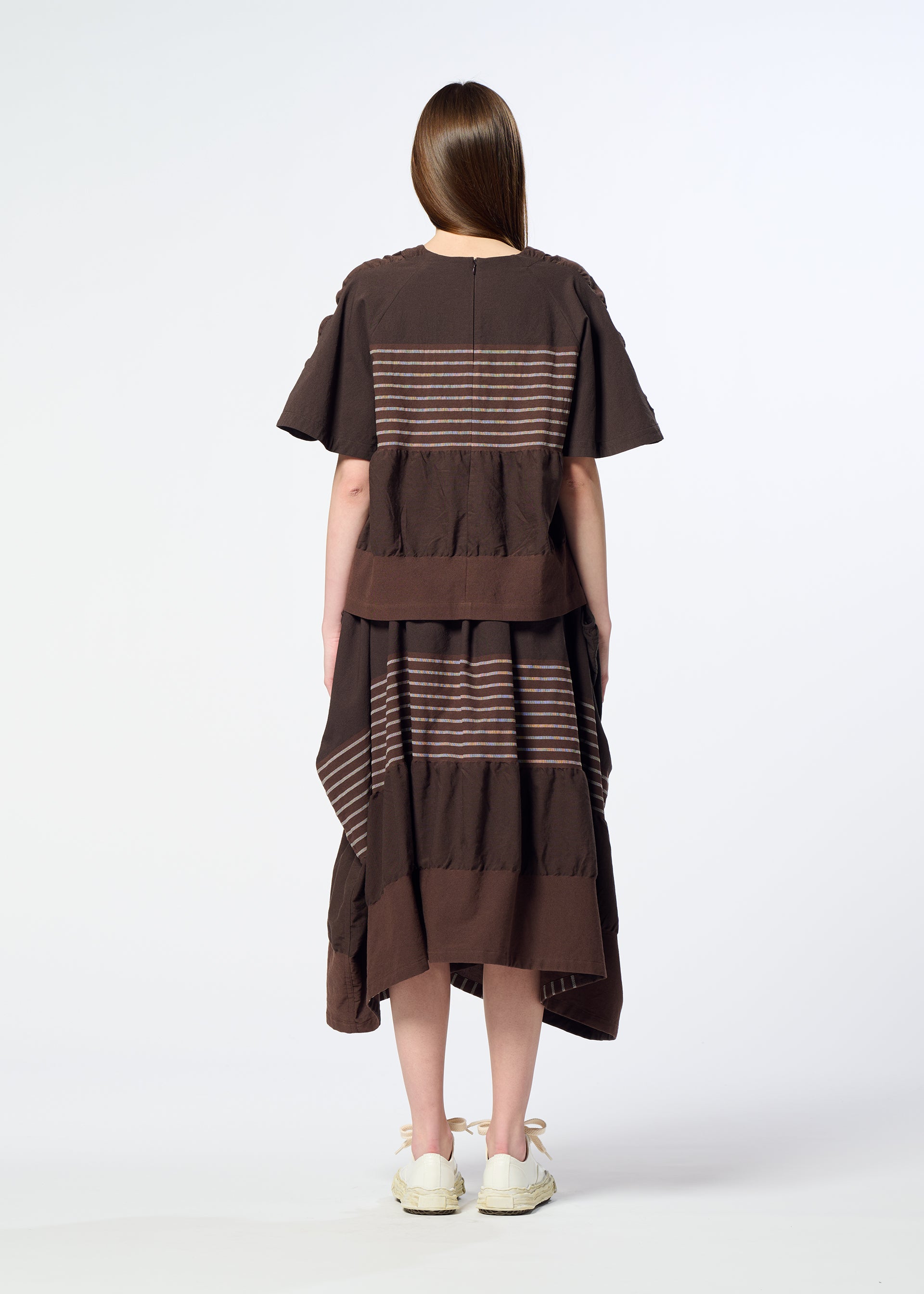 Zoomed view A person with long brown hair, dressed in HaaT’s COLLAGE STRETCH Ⅱ—a loose brown top paired with a voluminous skirt featuring horizontal colorful stripes—stands facing away from the camera against a plain white background.