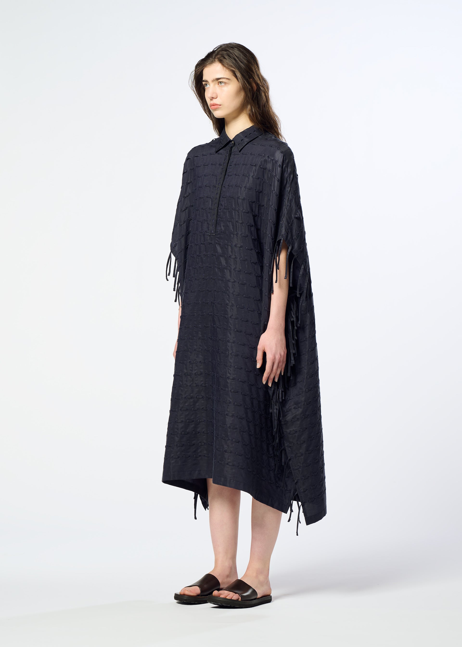 Zoomed view A person stands against a plain white background wearing the HaaT HOLE & LINE, a loose, textured black poncho-style dress with short sleeves and tassel details on the sleeves and sides, paired with black sandals.