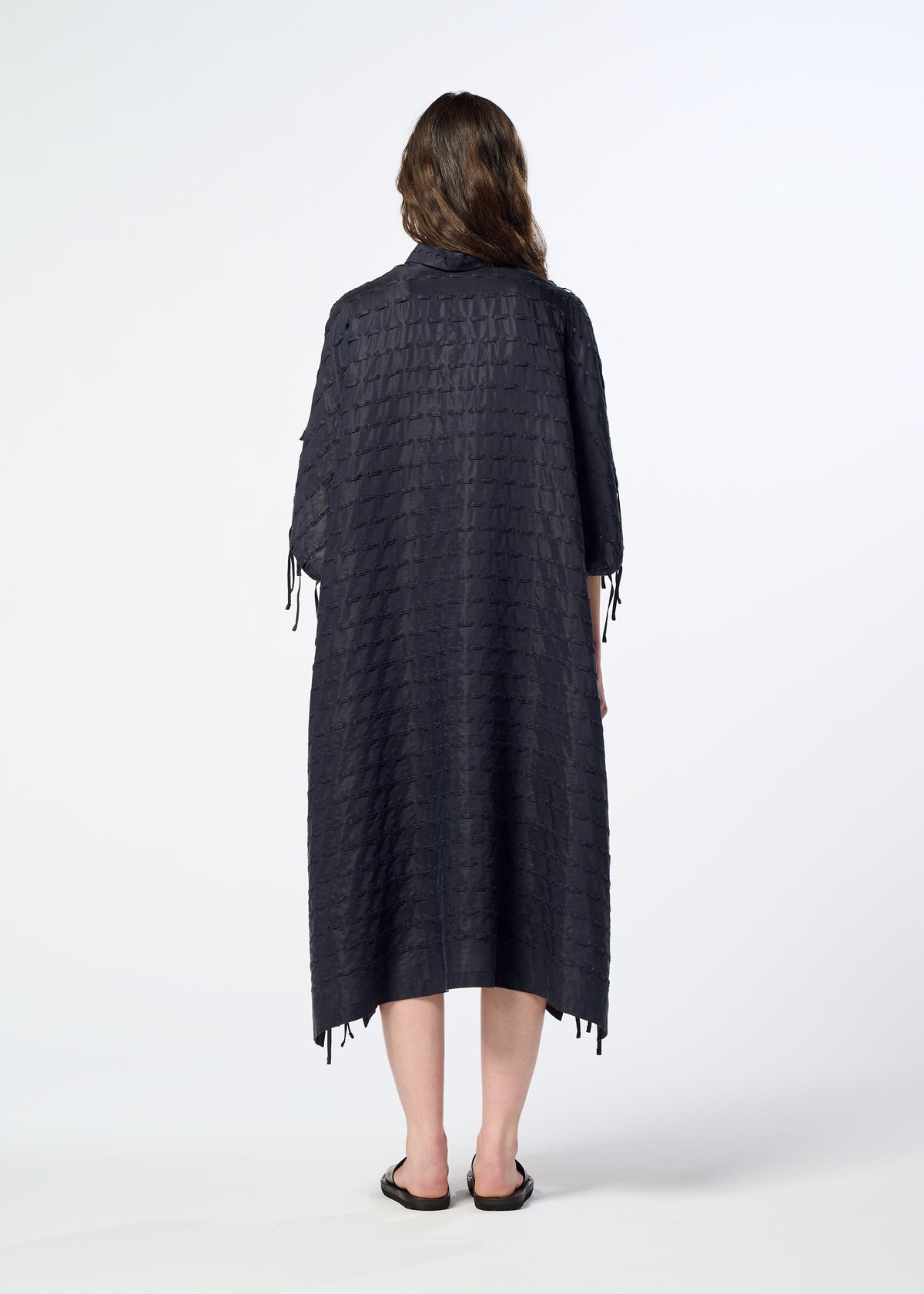 A person stands facing away against a plain white background, wearing HaaT’s HOLE & LINE textured, loose-fitting poncho-style dress with elbow-length sleeves and black sandals. view 8 of 8