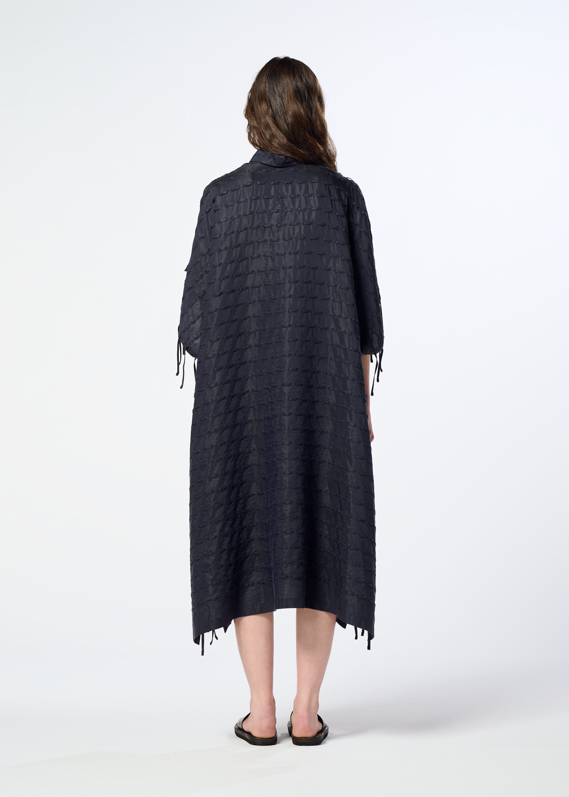 Zoomed view A person stands facing away against a plain white background, wearing HaaT’s HOLE & LINE textured, loose-fitting poncho-style dress with elbow-length sleeves and black sandals.