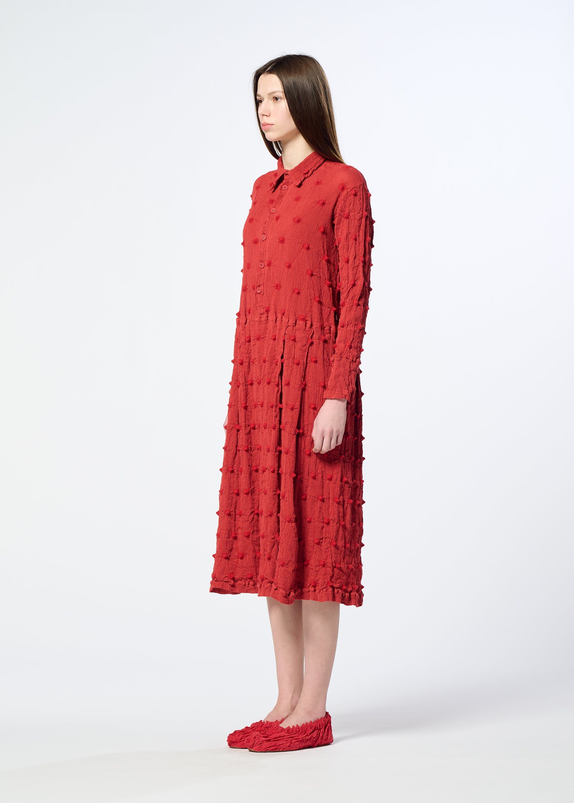 Zoomed view A woman stands in profile wearing the HaaT KYO CHIJIMI SQUARE EMB dress, which features a crinkled texture with long sleeves and a hemline that extends below the knee. She pairs the outfit with matching red shoes, standing against a plain white background.