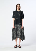 A woman with straight brown hair stands against a plain background, wearing HaaT’s ONE STROKE DRAW jacquard dress with white abstract lines and a handkerchief-hem skirt, paired with black sandals.