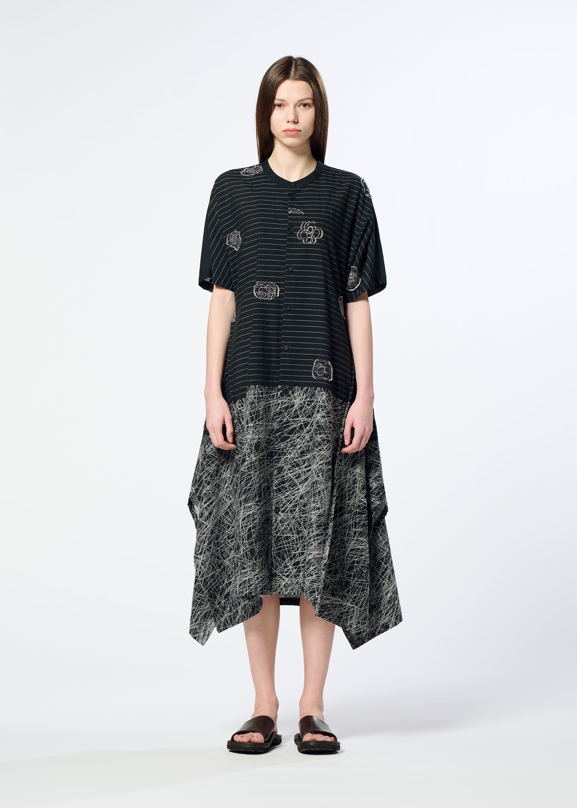 Zoomed view A woman with straight brown hair stands against a plain background, wearing HaaT’s ONE STROKE DRAW jacquard dress with white abstract lines and a handkerchief-hem skirt, paired with black sandals.