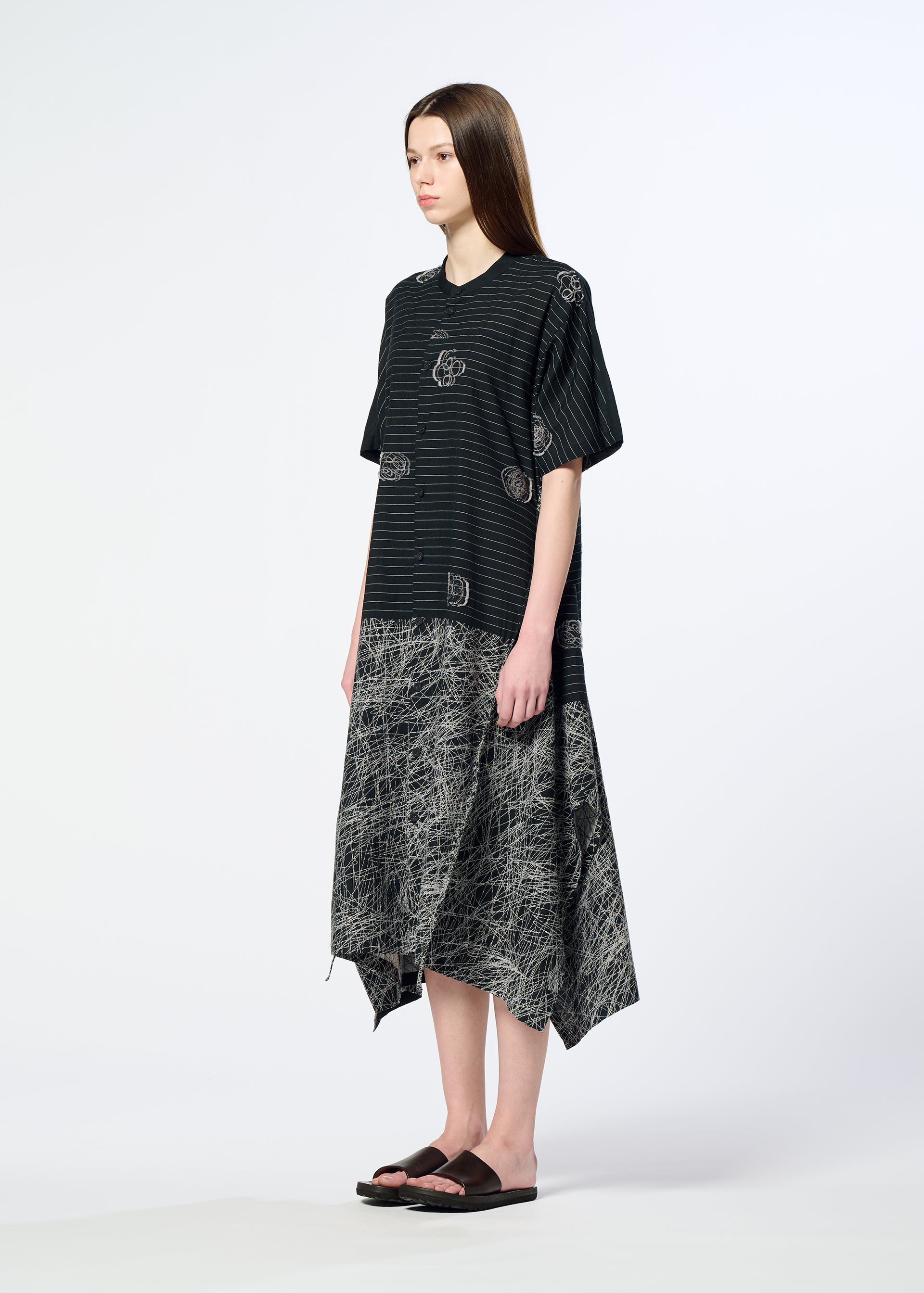 Zoomed view A woman with straight brown hair wears HaaT's ONE STROKE DRAW jacquard short-sleeved dress, featuring circular patterns and a black-and-gray asymmetrical textured skirt, paired with black sandals against a plain light background.