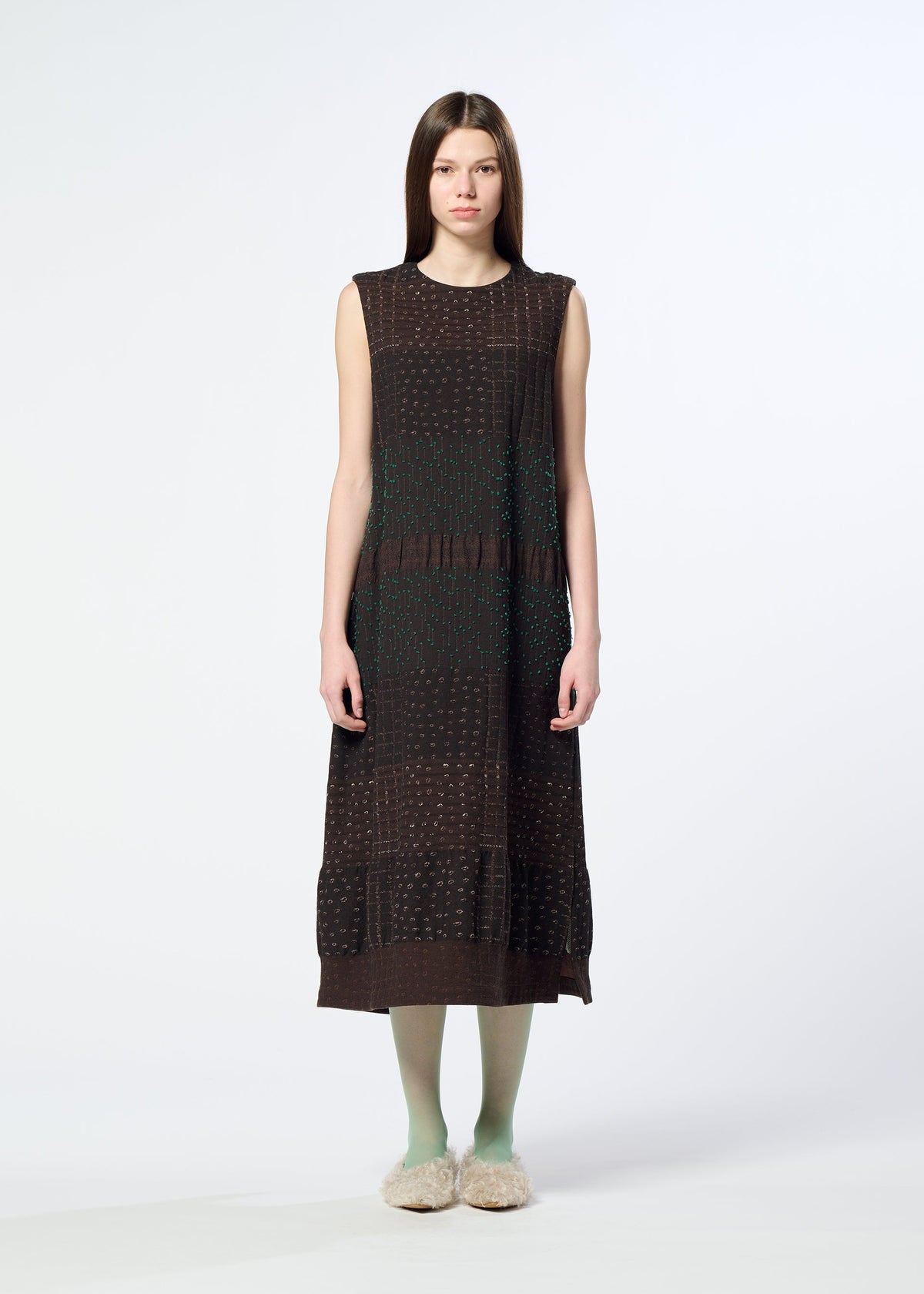 A woman stands facing forward against a plain white background, wearing the HaaT COLLAGE STRETCH Ⅰ sleeveless dark brown midi dress with a subtle check pattern, paired with light green tights and beige fuzzy slippers. view 6 of 8