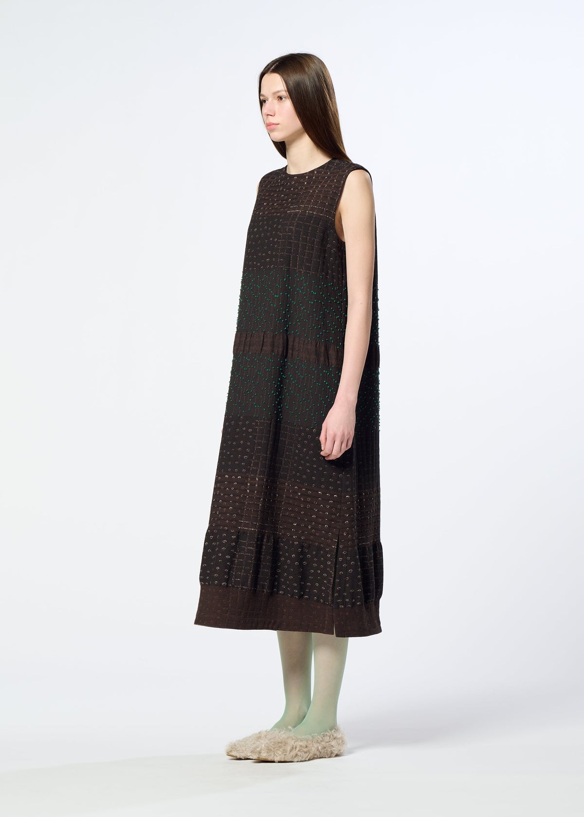 A woman with long straight hair wears the HaaT COLLAGE STRETCH Ⅰ, a loose, sleeveless dress in dark brown with subtle jacquard weaving. She pairs it with green tights and beige fuzzy slippers, standing against a plain white background and facing slightly to the side. view 7 of 8