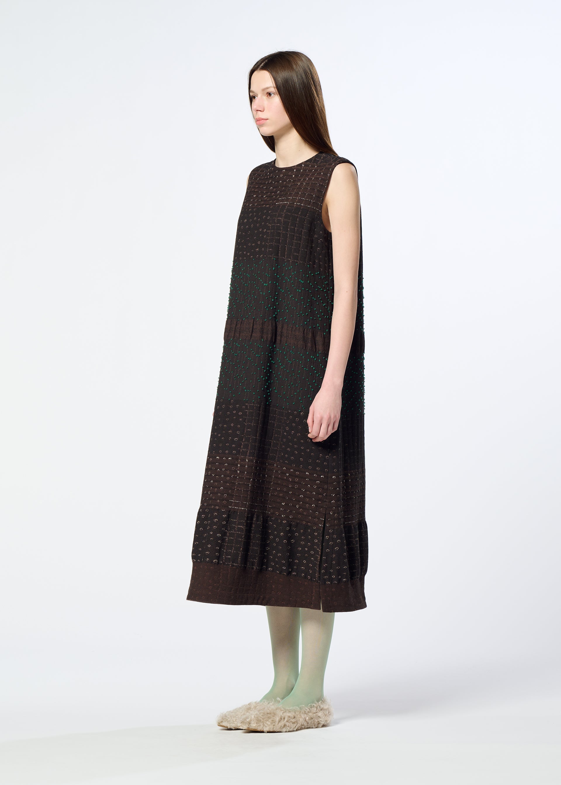 Zoomed view A woman with long straight hair wears the HaaT COLLAGE STRETCH Ⅰ, a loose, sleeveless dress in dark brown with subtle jacquard weaving. She pairs it with green tights and beige fuzzy slippers, standing against a plain white background and facing slightly to the side.