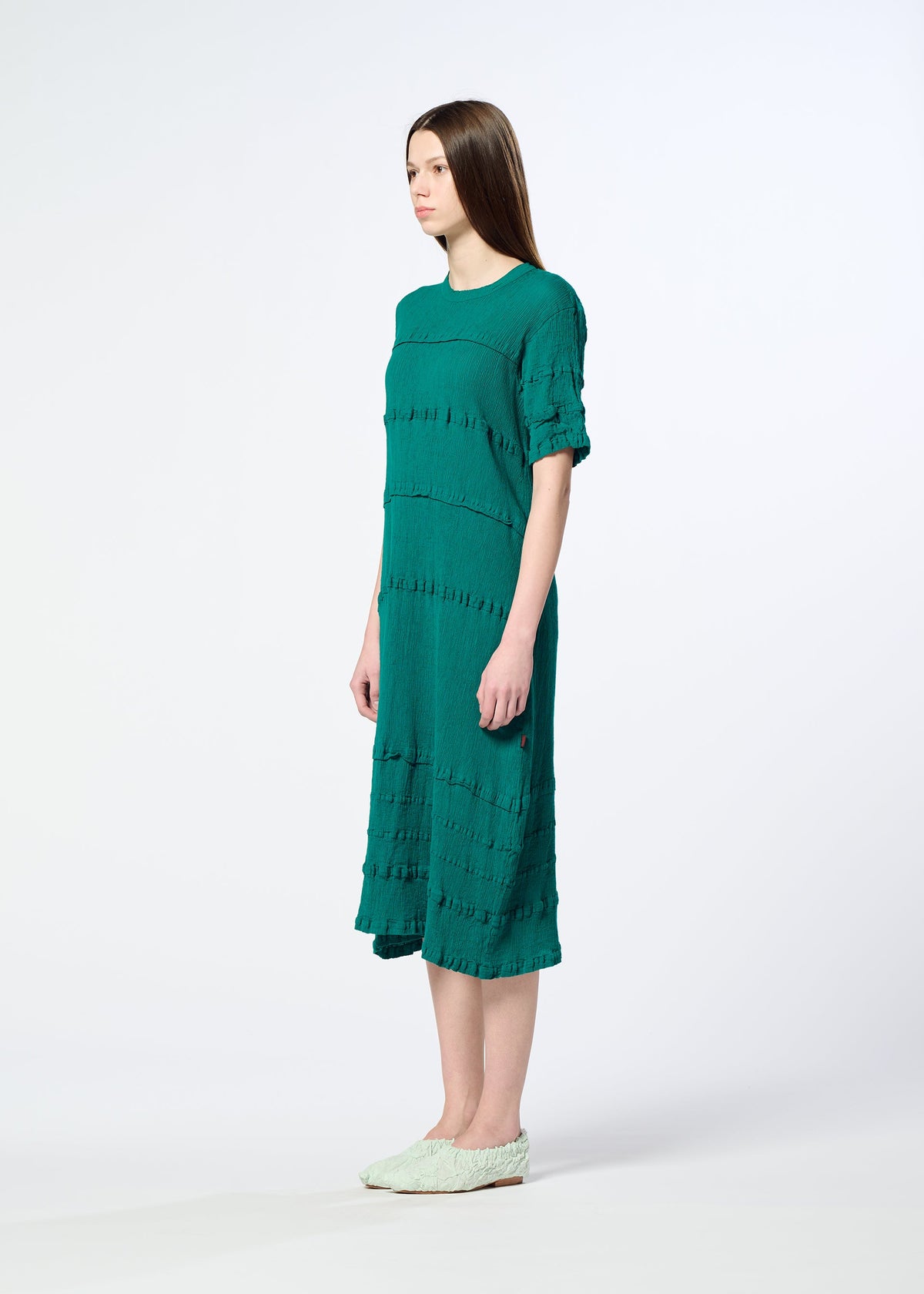 A woman stands sideways in a simple green KYO CHIJIMI TSUCHIKABE dress by HaaT with short sleeves and light green slip-on shoes, set against a plain white background. view 7 of 8