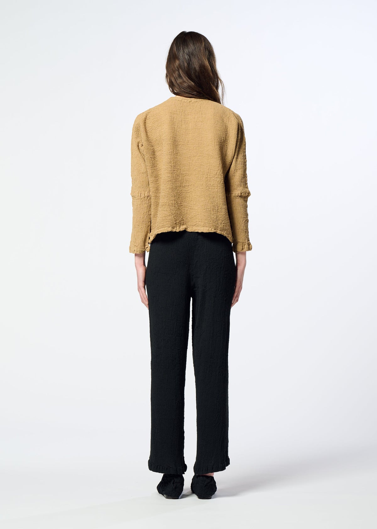 A person stands facing away, wearing a loose tan textured KYO CHIJIMI 2WAY top by HaaT and black pants, with long brown hair down. The round-neck long-sleeved top highlights the unique product dyeing process against a plain, light-colored background. view 8 of 8
