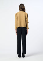 A person stands facing away, wearing a loose tan textured KYO CHIJIMI 2WAY top by HaaT and black pants, with long brown hair down. The round-neck long-sleeved top highlights the unique product dyeing process against a plain, light-colored background.