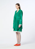 A woman in profile stands against a plain background, wearing HaaT's WALL WHITE HEM coat in green over a white dress with orange flats. Her straight hair is worn down, and the coat features subtle Kyoto dyeing for artistic elegance.