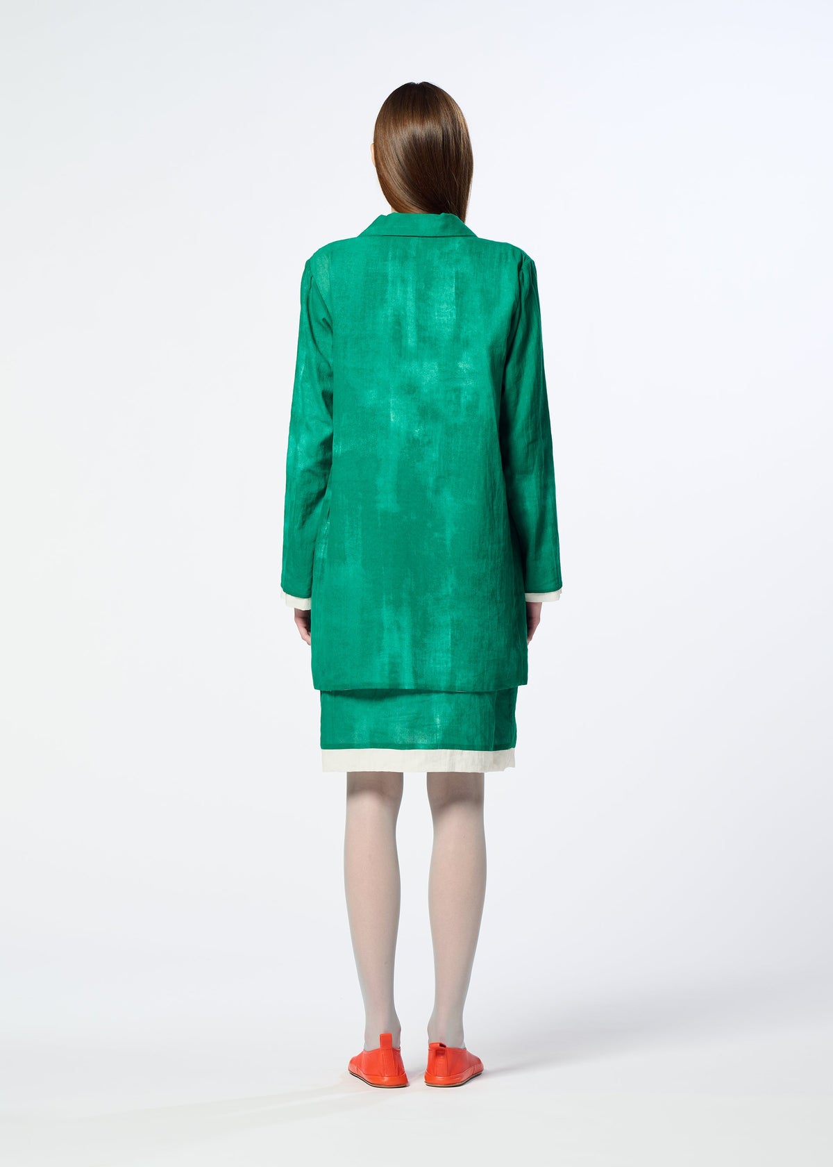 A person with straight brown hair, wearing HaaT's WALL WHITE HEM green long-sleeve jacket and skirt, light tights, bright orange flats, and a delicate Nassen print accent stands facing away on a white background. view 8 of 8