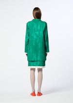 A person with straight brown hair, wearing HaaT's WALL WHITE HEM green long-sleeve jacket and skirt, light tights, bright orange flats, and a delicate Nassen print accent stands facing away on a white background.