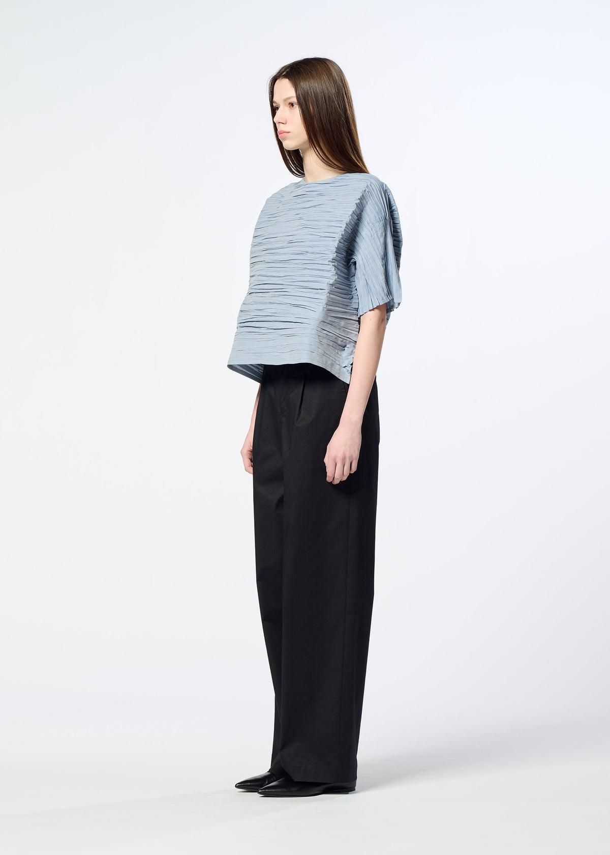 A woman stands sideways against a plain white background, wearing the HaaT SHIBARI SHIRT—a loose, light blue textured short-sleeved top with a subtle Shibari finish—paired with wide-leg black pants and black shoes. view 8 of 10