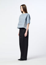 A woman stands sideways against a plain white background, wearing the HaaT SHIBARI SHIRT—a loose, light blue textured short-sleeved top with a subtle Shibari finish—paired with wide-leg black pants and black shoes.