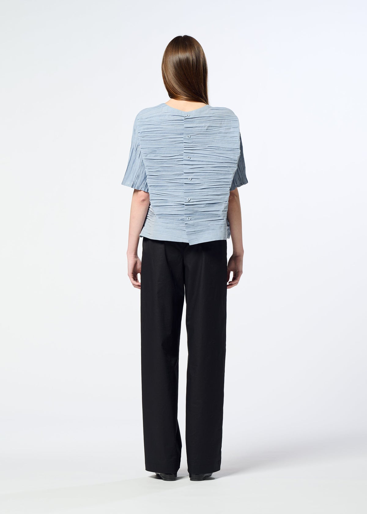 A person with long brown hair wears the HaaT SHIBARI SHIRT—a light blue, short-sleeved top featuring a Shibari finish and button details down the back, crafted from recycled polyester—paired with loose black pants. They stand against a white background, facing away from the camera. view 9 of 10