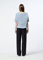 A person with long brown hair wears the HaaT SHIBARI SHIRT—a light blue, short-sleeved top featuring a Shibari finish and button details down the back, crafted from recycled polyester—paired with loose black pants. They stand against a white background, facing away from the camera.