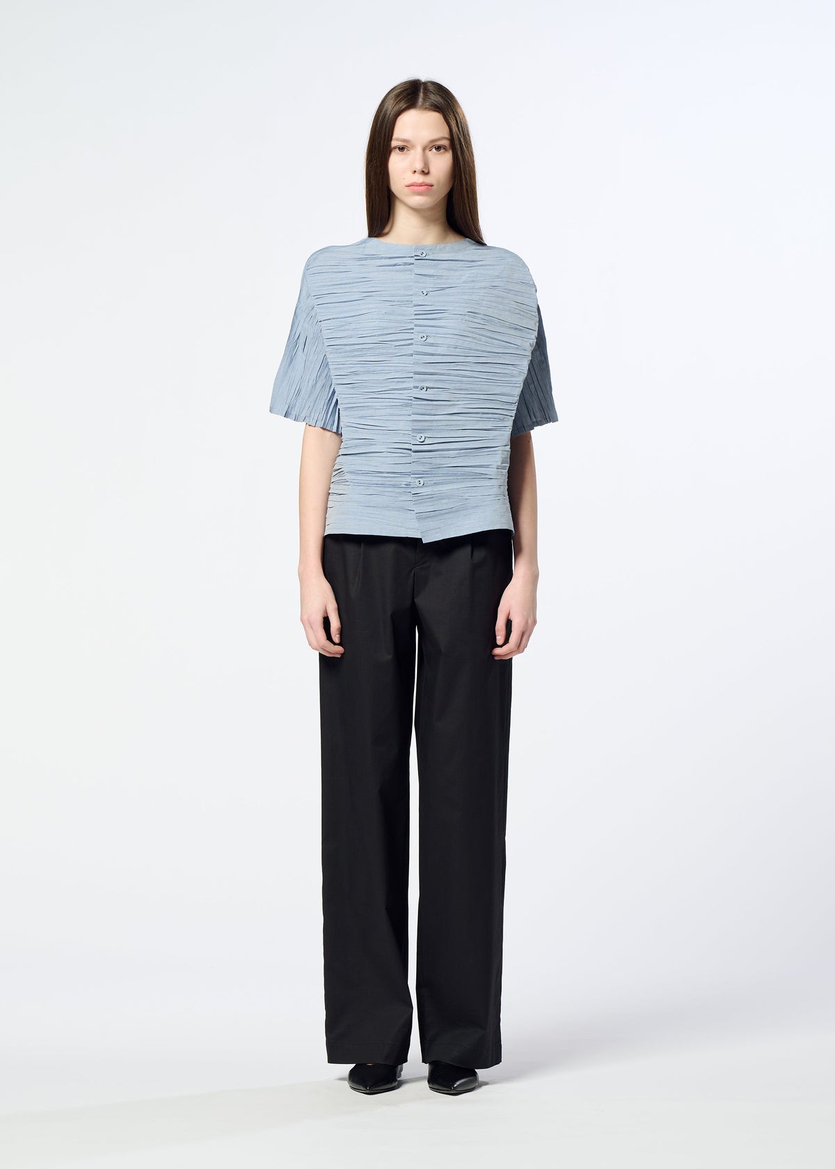 A person with long brown hair stands facing forward against a plain white background, wearing the HaaT SHIBARI SHIRT—a light blue textured short-sleeved top crafted from recycled polyester—paired with black wide-leg pants and black shoes. view 10 of 10