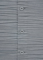 Close-up of the SHIBARI SHIRT by HaaT, a gray short-sleeved top featuring horizontal pleats and three buttons in the center. Crafted from recycled polyester for an eco-friendly touch, its soft texture and pleated arrangement create a wavy effect across the material.