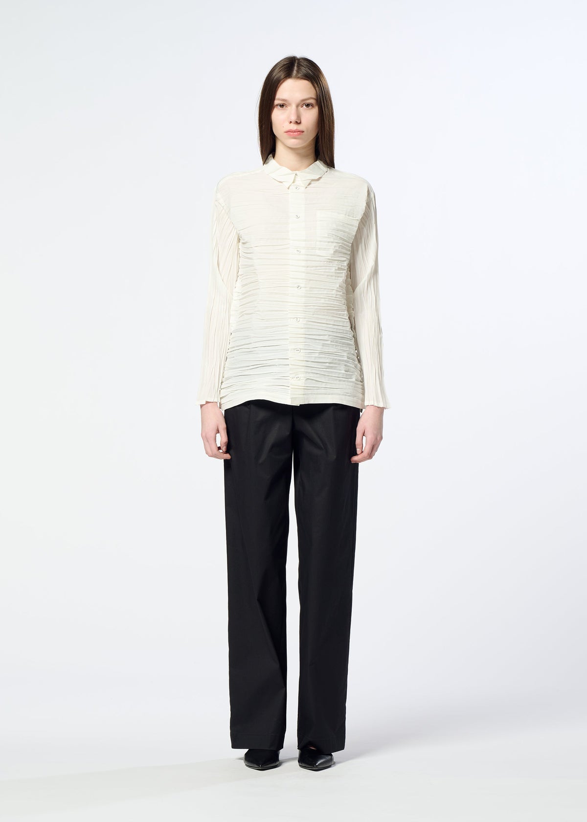 A woman stands facing forward against a plain white background, wearing the HaaT SHIBARI SHIRT— a cream, long-sleeved top featuring horizontal pleats and crafted from recycled polyester—paired with loose black trousers. She has straight brown hair and wears black shoes. view 7 of 9