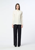 A woman stands facing forward against a plain white background, wearing the HaaT SHIBARI SHIRT— a cream, long-sleeved top featuring horizontal pleats and crafted from recycled polyester—paired with loose black trousers. She has straight brown hair and wears black shoes.
