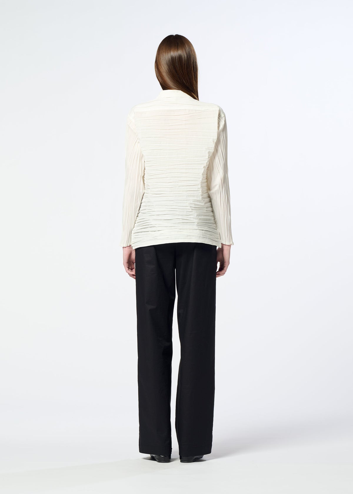 A person with straight brown hair stands facing away, wearing the SHIBARI SHIRT by HaaT—a long-sleeved, off-white textured blouse crafted from recycled polyester—paired with loose black pants against a plain light background. view 9 of 9