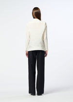 A person with straight brown hair stands facing away, wearing the SHIBARI SHIRT by HaaT—a long-sleeved, off-white textured blouse crafted from recycled polyester—paired with loose black pants against a plain light background.