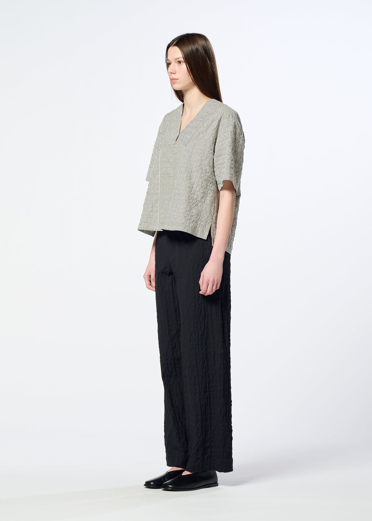 A woman with long straight hair stands sideways against a plain light background, wearing HaaT’s RIPPLE STRIPE loose-textured seersucker top paired with wide black pants and black shoes. view 6 of 7