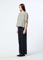 A woman with long straight hair stands sideways against a plain light background, wearing HaaT’s RIPPLE STRIPE loose-textured seersucker top paired with wide black pants and black shoes.